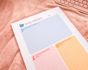 May include: A Daily Reflection worksheet with sections for gratitude, goals, and self-improvement. The worksheet has a light blue, peach, and orange color scheme with the words "Daily Reflection" in a cursive font. A pink keyboard is in the background.