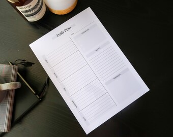 May include: A white daily plan notepad with the title "Daily Plan" is on a dark surface. The notepad has sections for time, priorities, notes, and reminders. A mug, a bottle, and a pair of glasses are also on the surface.
