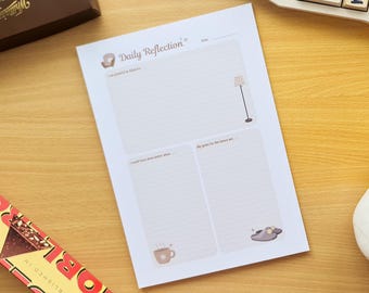 May include: A white Daily Reflection notepad with illustrations of a couch, lamp, coffee cup, and slippers. The notepad has prompts for gratitude, goals, and self-reflection. A chocolate bar and box are in the background.