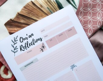 May include: A white journal page with the text "Qur'an Reflections" and floral illustrations. The page has sections for notes, dates, and verses, with a soft pink color scheme. The journal is set against a backdrop of patterned fabrics.
