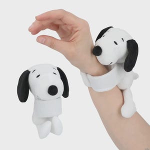 May include: Two plush Snoopy wristbands. The Snoopy figures are white with black ears and noses. One is worn on a wrist, the other is separate. The wristbands are designed to wrap around the wrist.