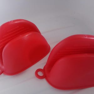 May include: A pair of bright red silicone pinch mitts featuring a curved, semi-circular shape with horizontal ribbed detailing on the top surface for grip and a small circular loop on the side for hanging storage. These kitchen tools are flexible.