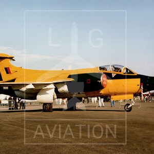 May include: A yellow and black military jet aircraft on the ground. The jet has a distinctive design with a pointed nose and a tail fin. The aircraft is parked on a paved surface with people in the background.