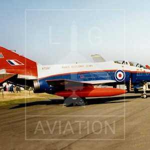 May include: A vintage military aircraft, primarily red, white, and blue, is parked on a tarmac. The tail features a red and white design with a blue and white emblem. The fuselage is white with blue accents and a roundel. The aircraft has a red fuel tank.