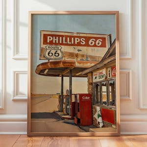 May include: Framed print of a vintage Phillips 66 gas station on Route 66. The sign displays "Phillips 66" and "Route 66". A red Coke machine and petrol pumps are visible. The artwork is in a wooden frame.