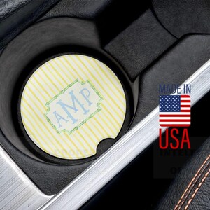 May include: A circular car cup holder coaster featuring a light yellow and white vertical striped pattern with a central light blue decorative frame containing the monogram letters AMP, positioned inside a black vehicle console.