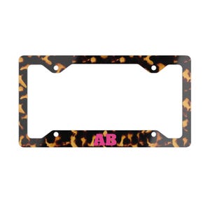 May include: A rectangular license plate frame featuring a brown and black tortoiseshell pattern. The frame has four circular mounting holes at the corners. Centered on the bottom edge of the frame are the bold, bright pink capital letters AB.