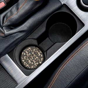 Könnte beinhalten: Ein runder Auto-Getränkehalter-Untersetzer mit braun-schwarzem Leopardenmuster. Der Untersetzer befindet sich in einem schwarzen Kunststoff-Getränkehalter der Mittelkonsole, neben einer schwarzen Lederschalthebelmanschette mit orangefarbenen Nähten.