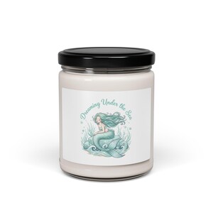 Pode incluir: Um frasco de vela branco com tampa preta. O rótulo apresenta uma ilustração de sereia turquesa com o texto "Dreaming Under the Sea". A sereia tem cabelo comprido turquesa e está sentada numa rocha no oceano.