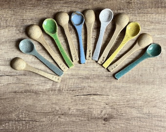 May include: A set of 12 ceramic spoons in various colors, arranged in a semi-circle on a wooden surface. The spoons are speckled with brown flecks and have a rounded bowl shape.