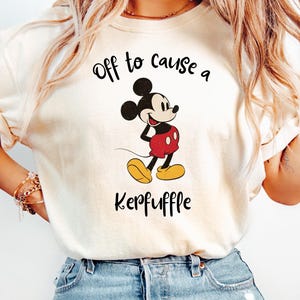 Könnte beinhalten: Elfenbeinfarbenes T-Shirt mit einer Mickey Mouse-Grafik in roter Hose und gelben Schuhen. Der Text "Off to cause a Kerfuffle" ist oberhalb und unterhalb der Figur in schwarzer Schrift gedruckt. Das T-Shirt wird mit einer blauen Jeans getragen.