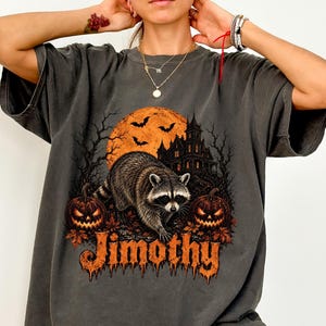 Könnte beinhalten: Ein anthrazitfarbenes T-Shirt mit einem Grafikdruck eines Waschbären vor einem großen orangefarbenen Mond, einem dunklen Herrenhaus und zwei Kürbislaternen. Das Wort Jimothy ist in einer stilisierten orangefarbenen Schrift unter dem Waschbären gedruckt.