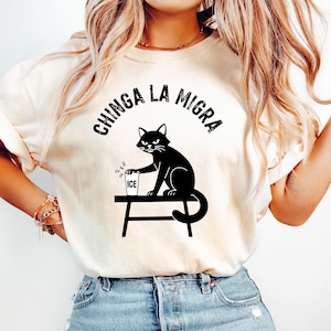 Könnte beinhalten: Cremefarbenes T-Shirt mit einer schwarzen Grafik einer Katze, die Eis in ein Glas gießt. Der Text "CHINGA LA MIGRA" ist über der Katze angeordnet. Die Katze sitzt auf einem kleinen Tisch.
