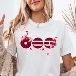 Könnte beinhalten: Ein weißes kurzärmeliges T-Shirt mit einem Grafikdesign aus drei kreisförmigen Formen in einer tiefroten, texturierten Optik. Das Motiv ist mit zarten rosa und roten Kirschblütenzweigen verziert, die sich um die kreisförmigen Elemente ranken.