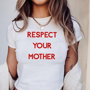 Könnte beinhalten: Ein weißes kurzärmeliges T-Shirt mit dem Text "RESPECT YOUR MOTHER", der in fetten roten Großbuchstaben auf der Brust gedruckt ist. Das Shirt wird mit einer goldfarbenen Kette und einer cremefarbenen Strickjacke über den Schultern kombiniert.