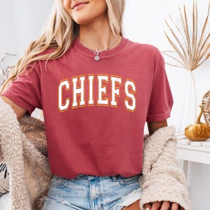 May include: A crimson short-sleeve t-shirt featuring the word CHIEFS printed in a distressed white font with a thin orange outline across the chest. The shirt has a crew neckline and is worn with light blue denim shorts and a fuzzy beige cardigan.