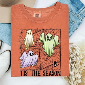 May include: A burnt orange t-shirt featuring a graphic of three ghosts in a spiderweb pattern with the text 'TIS' THE SEASON' printed at the bottom. The ghosts are coloured white, light green, and light purple, with black spider accents.