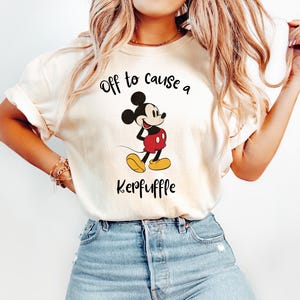 Könnte beinhalten: Cremefarbenes T-Shirt mit einer Grafik von Mickey Mouse in seiner klassischen Kleidung. Der Text "Off to cause a Keppuffle" ist über und unter der Figur aufgedruckt. Das Shirt wird mit einer blauen Jeans kombiniert.