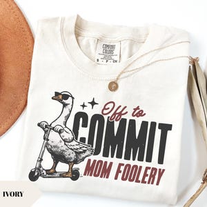Könnte beinhalten: Elfenbeinfarbenes T-Shirt mit einer Grafik einer Gans mit Sonnenbrille auf einem Roller. Der Text lautet "Off to COMMIT MOM FOOLERY". Eine goldene Halskette und ein brauner Hut sind ebenfalls zu sehen.