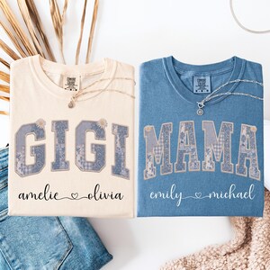 Könnte beinhalten: Zwei T-Shirts, eines cremefarben und eines blau, mit den Wörtern "GIGI" und "MAMA" in einem Patchwork-Design. Das cremefarbene Shirt hat die Namen "amelie" und "olivia" darunter, und das blaue Shirt hat "emily" und "michael". Jedes Shirt hat eine Halskette.