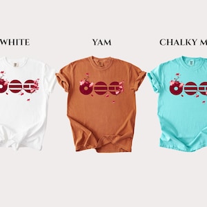 May include: Three short-sleeved t-shirts in white, yam, and chalky mint colors. Each shirt features a graphic of three dark red circles with horizontal lines and pink cherry blossom branches. The text labels WHITE, YAM, and CHALKY MINT appear above.