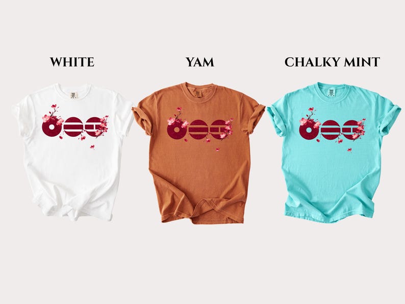 May include: Three short-sleeved t-shirts in white, yam, and chalky mint colors. Each shirt features a graphic of three dark red circles with horizontal lines and pink cherry blossom branches. The text labels WHITE, YAM, and CHALKY MINT appear above.