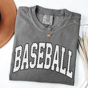 Könnte beinhalten: Ein graues Comfort Colors T-Shirt mit dem Wort "BASEBALL" in großen weißen, gepunkteten Buchstaben. Eine goldene Halskette mit Anhänger liegt auf dem Shirt. Das Shirt ist gefaltet, und ein brauner Hut ist teilweise im Hintergrund sichtbar.