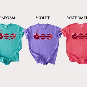 May include: Three short-sleeve crew neck t-shirts in seafoam, violet, and watermelon colors. Each shirt features a graphic design of three dark red circles with horizontal lines and pink cherry blossom branches extending from the right side.