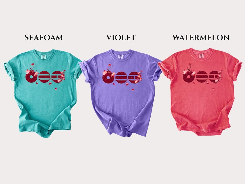 May include: Three short-sleeve crew neck t-shirts in seafoam, violet, and watermelon colors. Each shirt features a graphic design of three dark red circles with horizontal lines and pink cherry blossom branches extending from the right side.