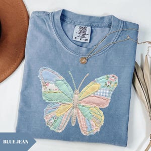 Könnte beinhalten: Ein blaues Comfort Colors T-Shirt mit einem Patchwork-Schmetterlingsdesign in Pastellfarben. Der Schmetterling hat rosa, gelbe, grüne und blaue Abschnitte. Eine goldene Halskette mit Anhänger liegt auf dem Shirt. Größe S.