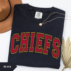 May include: A black short-sleeved t-shirt featuring the word CHIEFS in a large, arched, distressed red font with a yellow outline. The shirt has a Comfort Colors brand label at the inner neckline and is displayed with a gold-toned pendant necklace.