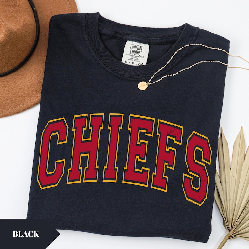 May include: A black short-sleeved t-shirt featuring the word CHIEFS in a large, arched, distressed red font with a yellow outline. The shirt has a Comfort Colors brand label at the inner neckline and is displayed with a gold-toned pendant necklace.