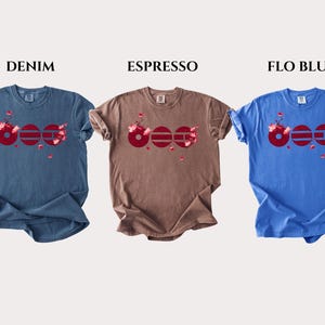 May include: Three short-sleeve t-shirts in denim blue, espresso brown, and flo blue. Each shirt features a graphic of three dark red circles with horizontal lines and pink cherry blossom accents. The text DENIM, ESPRESSO, and FLO BLUE appears above.