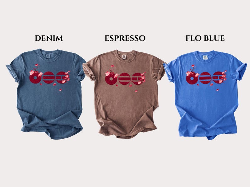 May include: Three short-sleeve t-shirts in denim blue, espresso brown, and flo blue. Each shirt features a graphic of three dark red circles with horizontal lines and pink cherry blossom accents. The text DENIM, ESPRESSO, and FLO BLUE appears above.