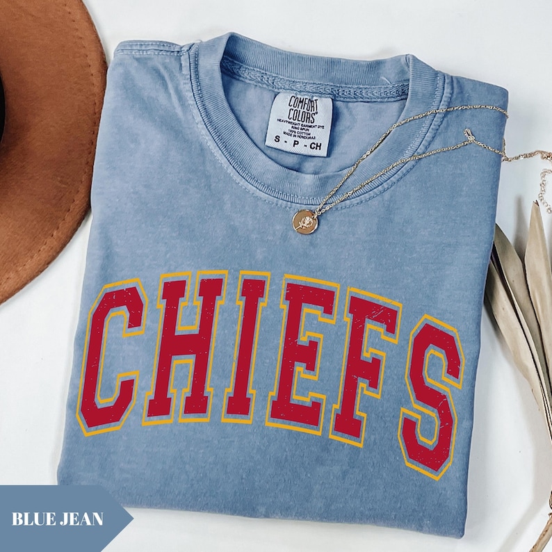 May include: A blue short-sleeved t-shirt with a crew neckline and the word CHIEFS printed in red with a yellow outline across the chest. The shirt features a Comfort Colors brand label at the neck and is paired with a gold-toned pendant necklace.