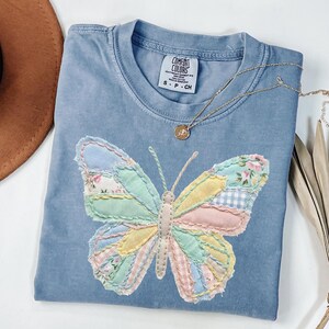 Könnte beinhalten: Hellblaues Comfort Colors T-Shirt mit einem Patchwork-Schmetterlingsdesign in Pastellfarben. Der Schmetterling hat Flügel mit floralen, Vichy- und einfarbigen Stoffflicken. Eine goldene Halskette mit Anhänger liegt auf dem Shirt.