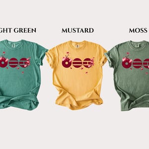 May include: Three short-sleeve t-shirts in light green, mustard, and moss colors. Each shirt features a graphic of the word "BOO" in dark red with cherry blossom branches and pink flowers integrated into the letters, labeled LIGHT GREEN, MUSTARD, MOSS.