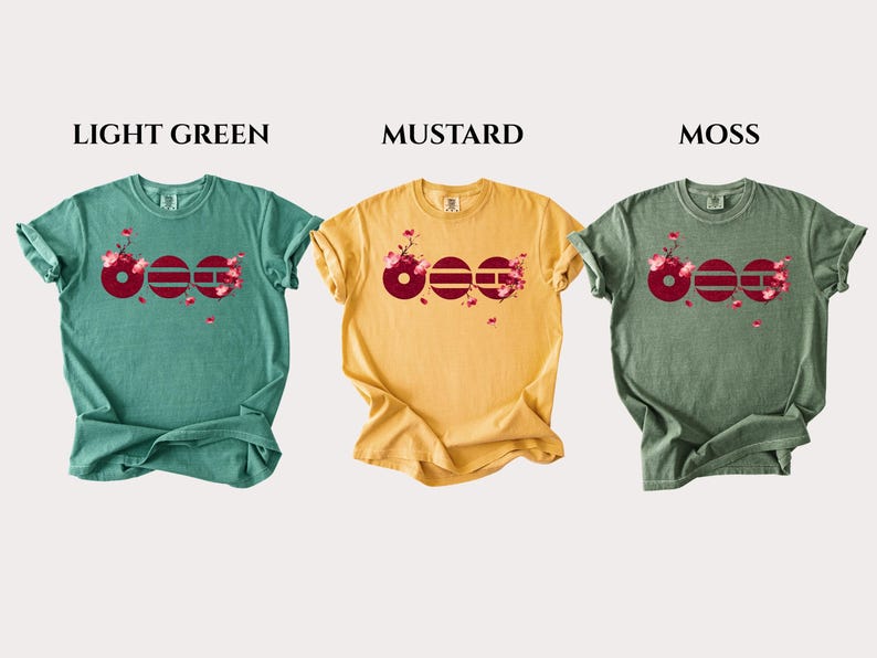May include: Three short-sleeve t-shirts in light green, mustard, and moss colors. Each shirt features a graphic of the word "BOO" in dark red with cherry blossom branches and pink flowers integrated into the letters, labeled LIGHT GREEN, MUSTARD, MOSS.