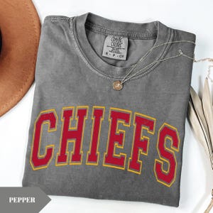 May include: A charcoal gray short-sleeve t-shirt with a crew neckline and a Comfort Colors brand label. The shirt features the word CHIEFS printed in a large, arched, red font with a yellow outline. A thin silver chain necklace rests on the fabric.