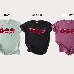 May include: Three short-sleeve t-shirts in bay, black, and berry colors. Each shirt features a graphic of three dark red circles with horizontal lines and pink cherry blossom branches. The words BAY, BLACK, and BERRY are printed above each shirt.