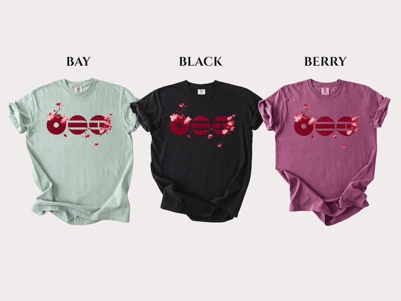 May include: Three short-sleeve t-shirts in bay, black, and berry colors. Each shirt features a graphic of three dark red circles with horizontal lines and pink cherry blossom branches. The words BAY, BLACK, and BERRY are printed above each shirt.