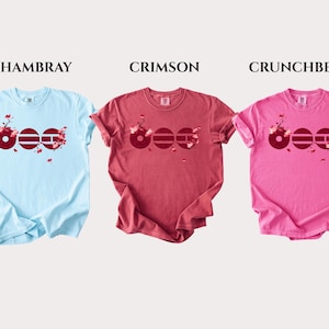 May include: Three short-sleeve t-shirts in light blue, crimson, and pink. Each shirt features a graphic of three dark red circles with horizontal lines and cherry blossom branches. The text labels CHAMBRAY, CRIMSON, and CRUNCHBERRY appear above.