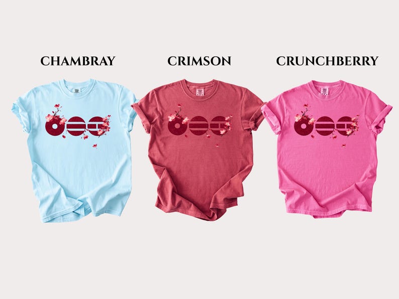 May include: Three short-sleeve t-shirts in light blue, crimson, and pink. Each shirt features a graphic of three dark red circles with horizontal lines and cherry blossom branches. The text labels CHAMBRAY, CRIMSON, and CRUNCHBERRY appear above.
