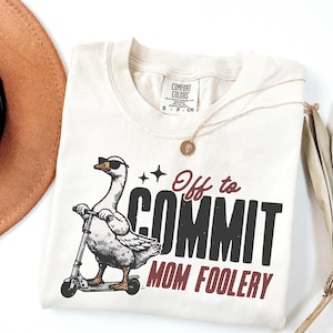 Könnte beinhalten: Cremefarbenes T-Shirt mit einer Grafik einer Gans mit Sonnenbrille auf einem Roller. Der Text lautet "Off to COMMIT MOM FOOLERY" in Schwarz und Burgunderrot. Eine goldene Halskette und ein brauner Hut sind ebenfalls zu sehen.