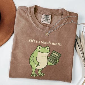 Könnte beinhalten: Ein braunes T-Shirt mit dem Text "Off to teach math" und einem Cartoon-Frosch, der einen Taschenrechner hält. Das Shirt hat einen Rundhalsausschnitt und kurze Ärmel. Eine silberne Halskette mit Anhänger liegt auf dem Shirt.