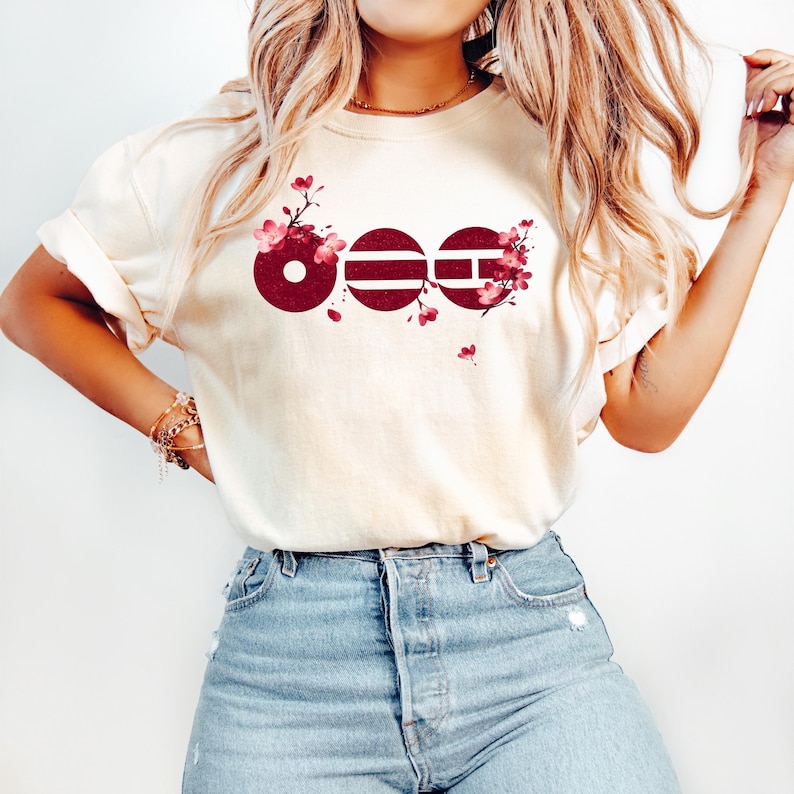 May include: A cream-colored short-sleeve t-shirt featuring a graphic of three dark red circular shapes with horizontal lines, decorated with pink cherry blossom branches. The shirt is paired with light blue denim jeans with a button fly closure.