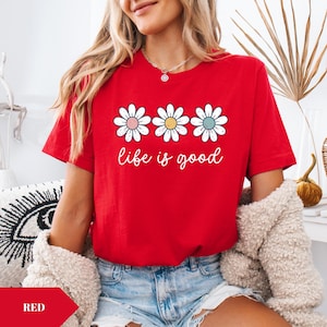 以下が含まれることがあります： パステルカラーのセンターを持つ3つのデイジーの花と、白いスクリプトで「life is good」というフレーズが特徴の赤いTシャツ。 このシャツは半袖で、柔らかい素材で作られています。