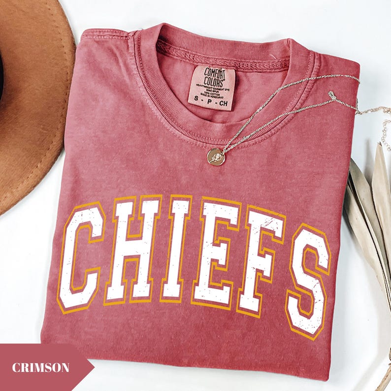 May include: A crimson short-sleeve t-shirt with a distressed white and yellow graphic that reads CHIEFS in a collegiate font. The shirt features a Comfort Colors brand label at the inner neckline and is paired with a delicate silver-toned necklace.