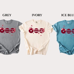 May include: Three short-sleeve crew neck t-shirts in grey, ivory, and ice blue. Each shirt features a graphic design of three dark red circles with horizontal lines, accented by pink cherry blossom branches and falling petals across the chest.