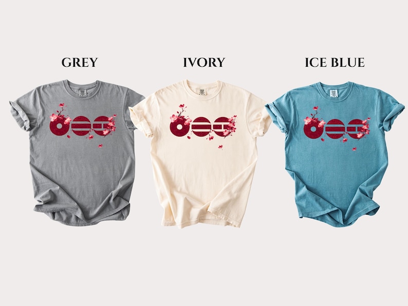 May include: Three short-sleeve crew neck t-shirts in grey, ivory, and ice blue. Each shirt features a graphic design of three dark red circles with horizontal lines, accented by pink cherry blossom branches and falling petals across the chest.